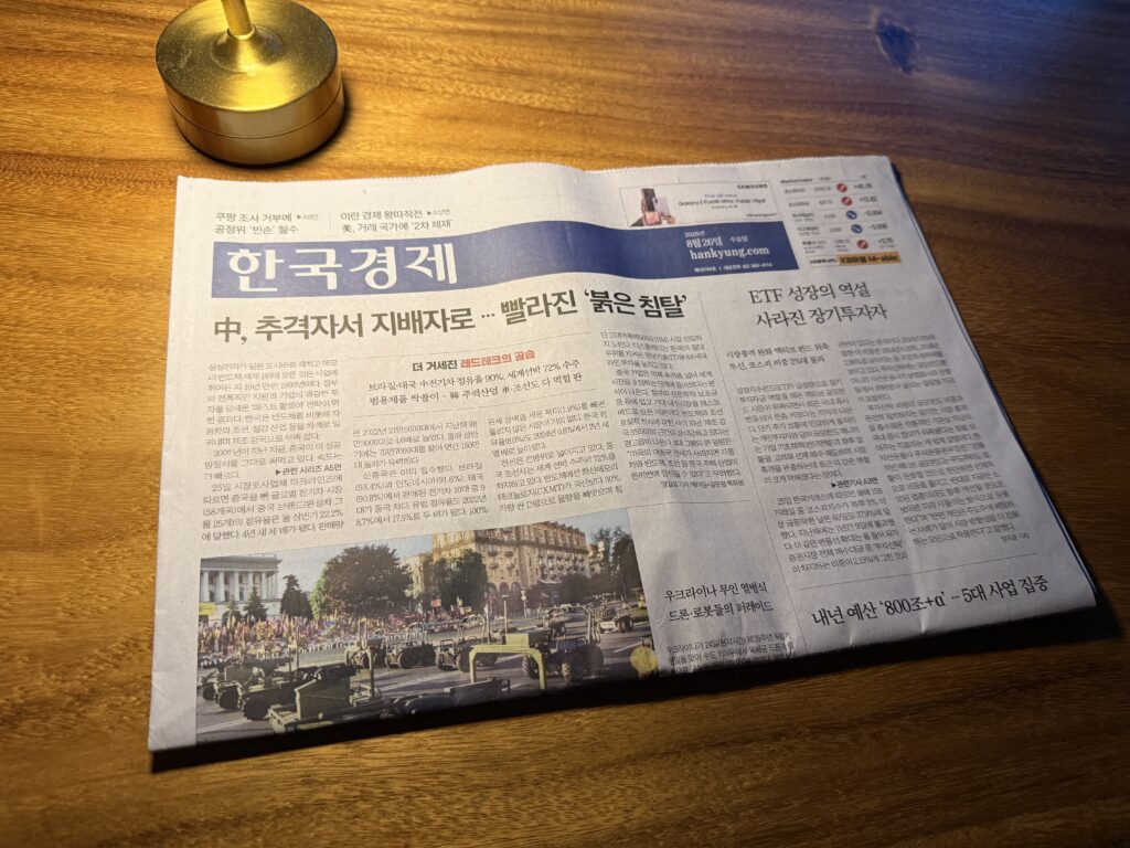 어두운 조명 아래 원목 식탁에 펼쳐 둔 한국경제신문 1면, 위쪽에 놋쇠 촛대가 놓여 있다. 머리기사는 '中, 추격자서 지배자로 … 빨라진 붉은 침탈'이고 아래에는 광장에서 군용 차량이 줄지어 지나가는 열병식 사진이 실려 있다.