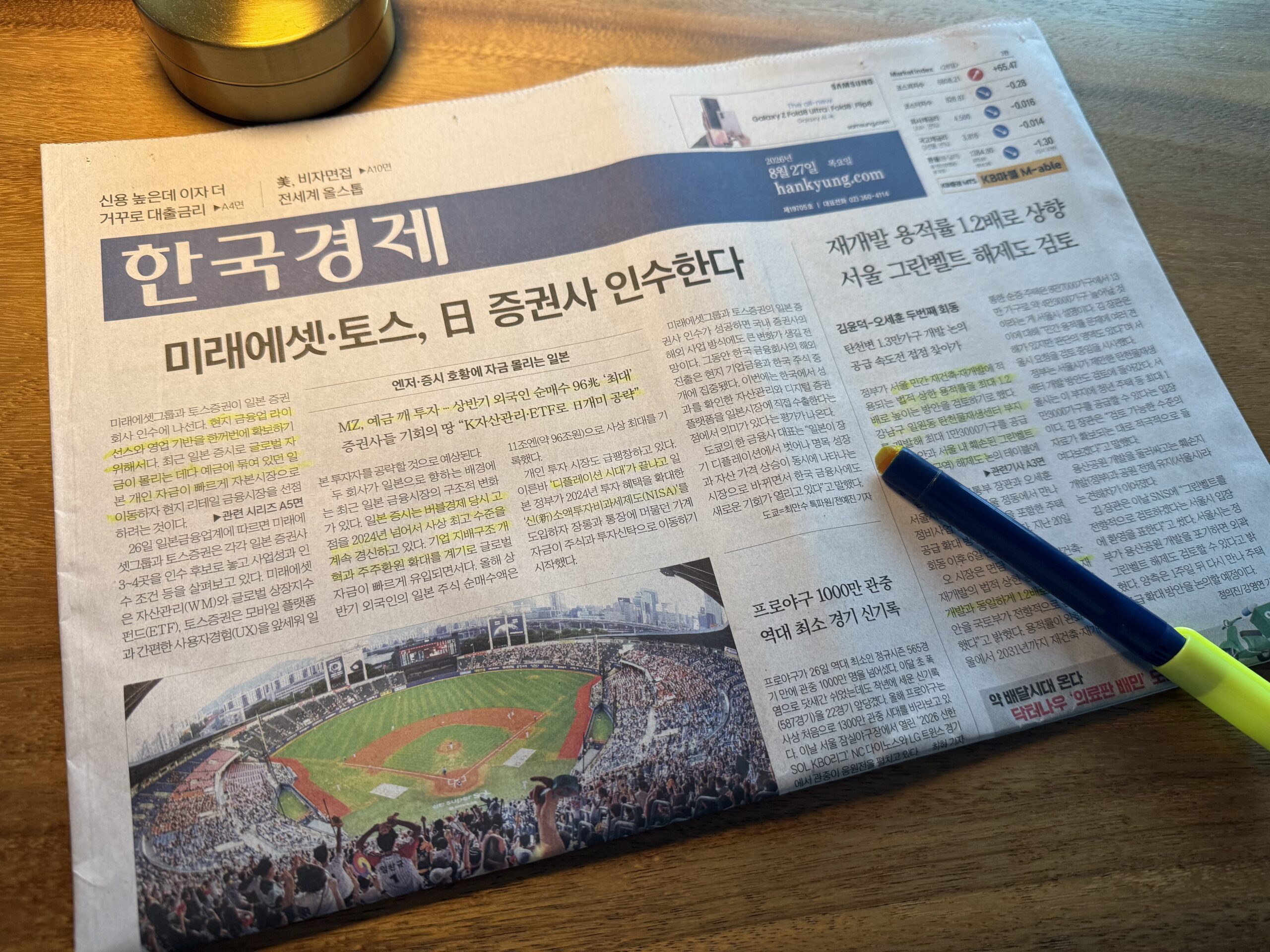 나무 책상 위에 펼쳐 둔 한국경제신문 1면과 볼펜·형광펜. 머리기사는 '미래에셋·토스, 日 증권사 인수한다'이고 왼쪽 첫 문단에 형광펜이 그어져 있다. 아래에는 관중이 가득 찬 야구장을 위에서 내려다보며 찍은 사진이 실려 있다.