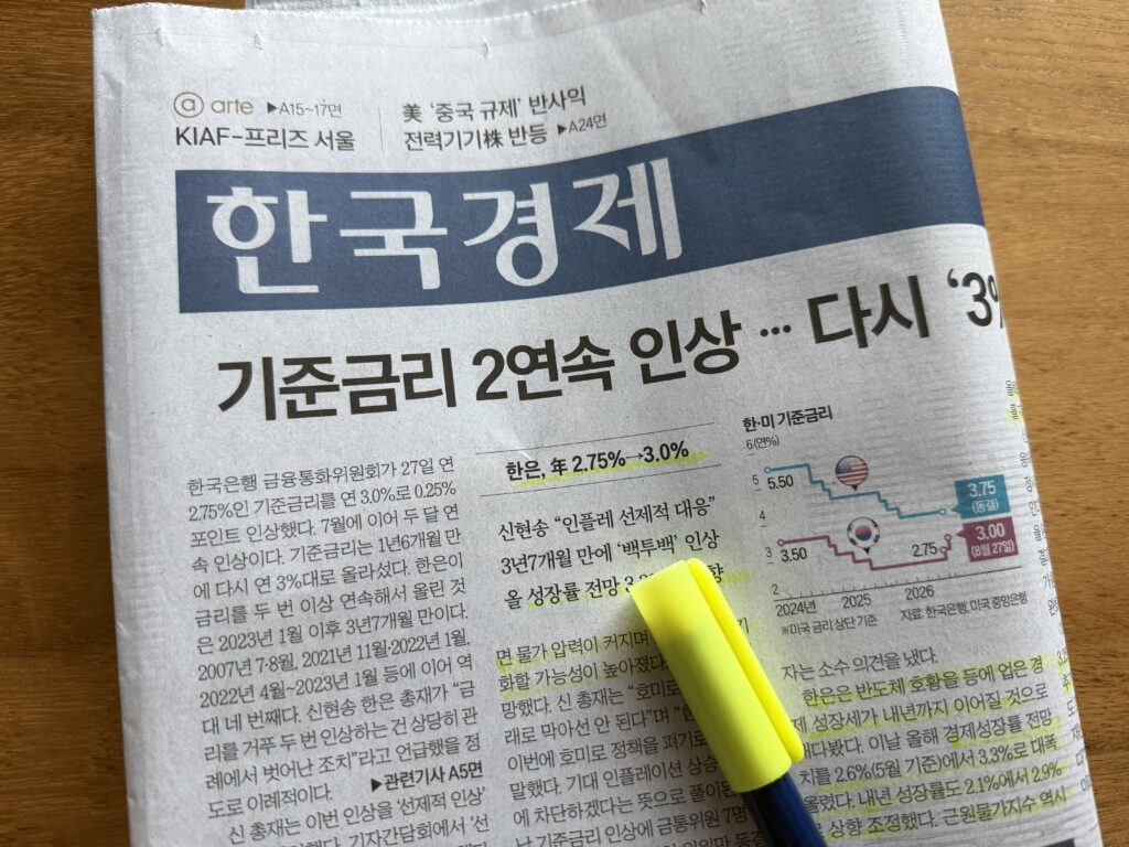 나무 책상 위 한국경제신문 1면과 노란 형광펜. 머리기사 '기준금리 2연속 인상 … 다시 3% 시대' 아래 본문 여러 줄에 형광펜이 그어져 있고, 오른쪽에는 한국과 미국 기준금리가 계단처럼 엇갈리며 움직인 꺾은선 그래프가 실려 있다.
