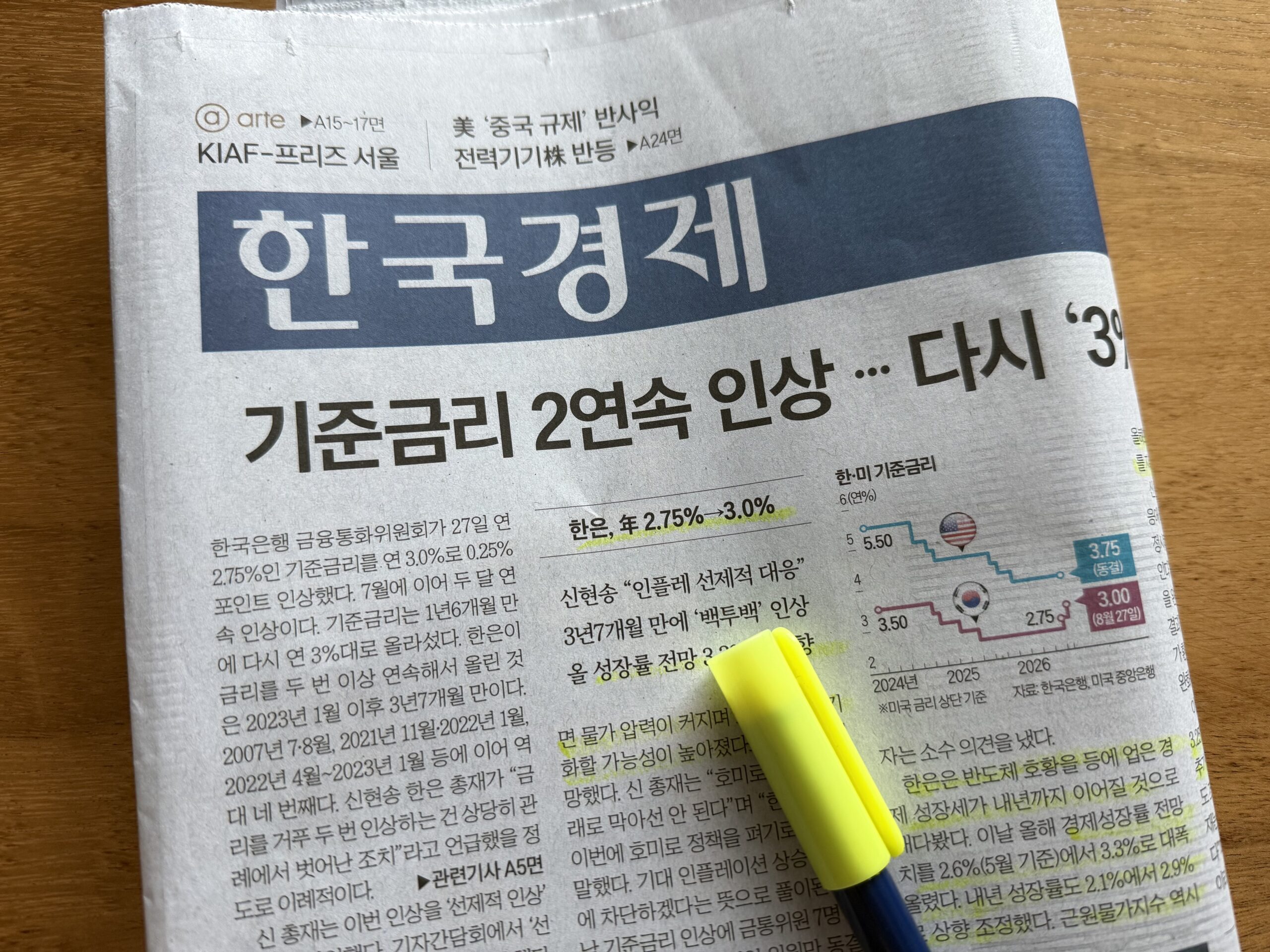 나무 책상 위 한국경제신문 1면과 노란 형광펜. 머리기사 '기준금리 2연속 인상 … 다시 3% 시대' 아래 본문 여러 줄에 형광펜이 그어져 있고, 오른쪽에는 한국과 미국 기준금리가 계단처럼 엇갈리며 움직인 꺾은선 그래프가 실려 있다.