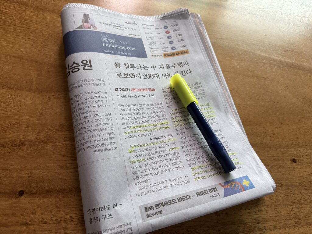 나무 책상 위에 펼쳐 둔 한국경제신문 지면과 형광펜. 위쪽에 '2026년 8월 31일 월요일 hankyung.com' 제호와 주요 지표 표가 보이고, 머리기사 '韓 침투하는 中 자율주행차 로보택시 200대 서울 달린다' 아래 본문 여러 곳에 형광펜이 그어져 있다.