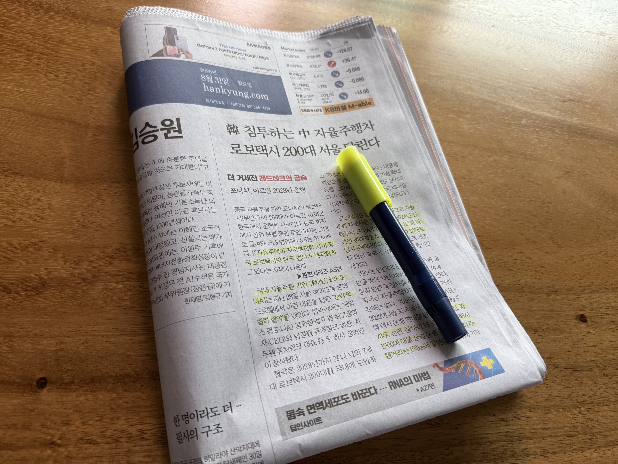 나무 책상 위에 펼쳐 둔 한국경제신문 지면과 형광펜. 위쪽에 '2026년 8월 31일 월요일 hankyung.com' 제호와 주요 지표 표가 보이고, 머리기사 '韓 침투하는 中 자율주행차 로보택시 200대 서울 달린다' 아래 본문 여러 곳에 형광펜이 그어져 있다.