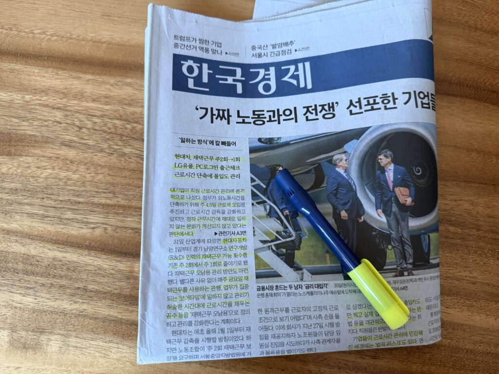 나무 책상 위 한국경제신문 1면과 볼펜·형광펜. 머리기사는 "'가짜 노동과의 전쟁' 선포한 기업들"이고 왼쪽 문단 여러 줄에 형광펜이 그어져 있다. 오른쪽에는 비행기 트랩에서 내려오는 남성들을 찍은 사진이 있다.