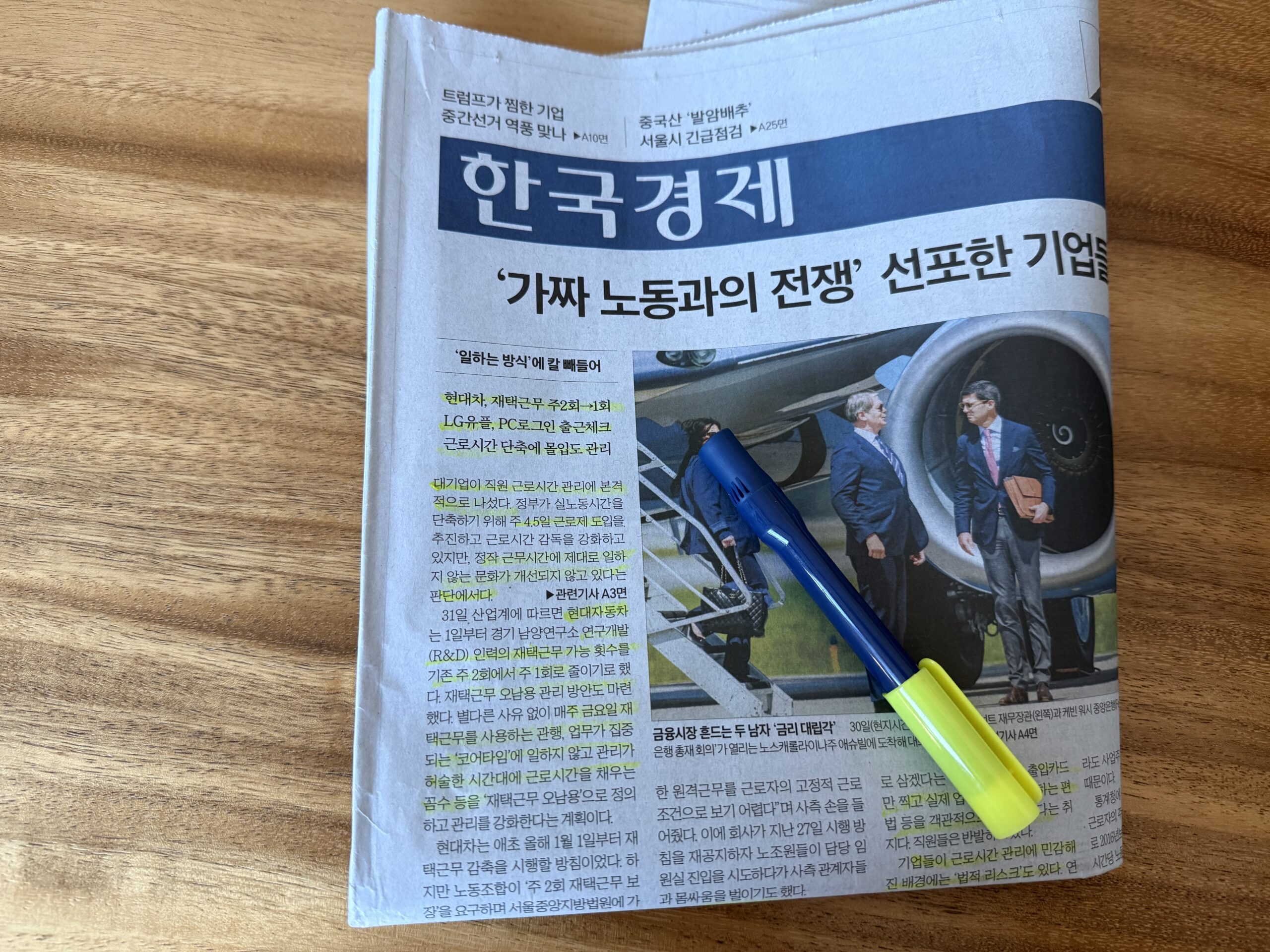 나무 책상 위 한국경제신문 1면과 볼펜·형광펜. 머리기사는 "'가짜 노동과의 전쟁' 선포한 기업들"이고 왼쪽 문단 여러 줄에 형광펜이 그어져 있다. 오른쪽에는 비행기 트랩에서 내려오는 남성들을 찍은 사진이 있다.