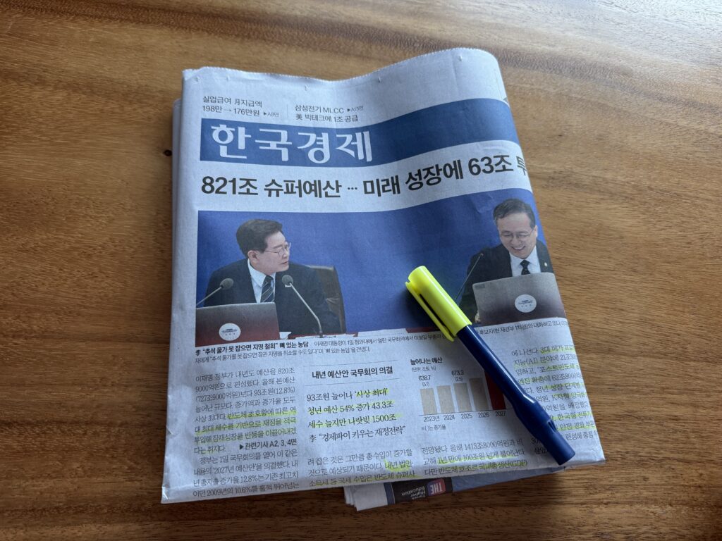 나무 책상 위 한국경제신문 1면과 노란 형광펜. 머리기사는 '821조 슈퍼예산 … 미래 성장에 63조'이고, 가운데에는 국무회의에서 마주 보고 이야기하는 두 인사의 사진, 아래에는 내년 예산이 늘어나는 막대그래프가 실려 있다.