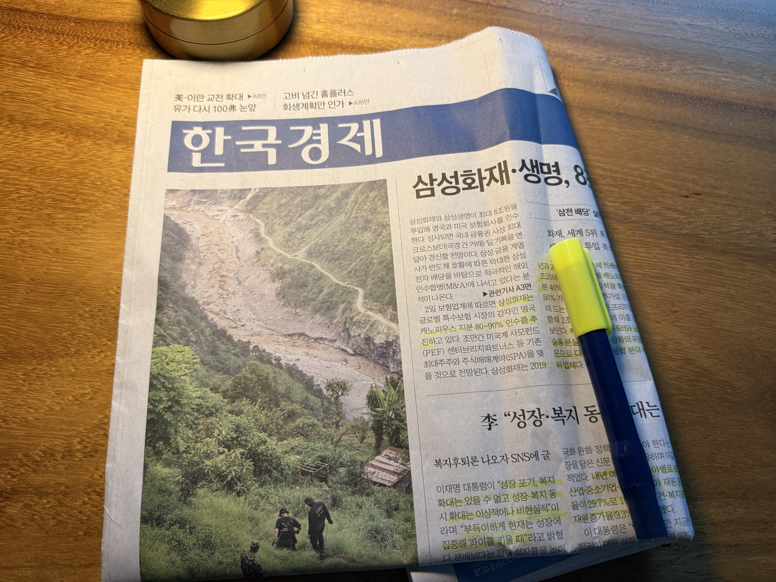 원목 책상 위에 놓인 한국경제신문 1면과 볼펜·형광펜, 위쪽에 금색 캔들 용기가 보인다. 머리기사는 '삼성화재·생명, 8조'이고 왼쪽에는 산사태로 흙이 쓸려 내려간 계곡을 사람들이 살펴보는 큰 사진이 실려 있다.