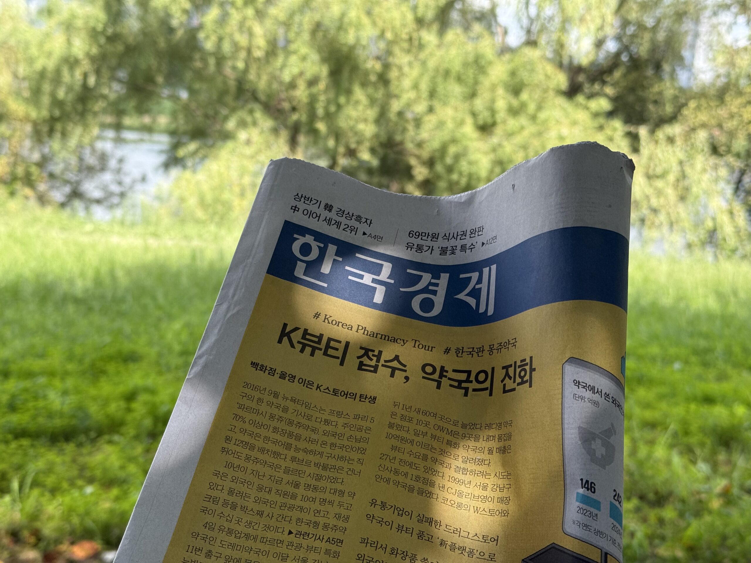잔디밭과 버드나무를 배경으로 손에 들고 펼친 한국경제신문 1면. 머리기사는 'K뷰티 접수, 약국의 진화'이고, 신문 뒤로 강가의 초록 잔디와 늘어진 나뭇가지가 흐릿하게 보인다.