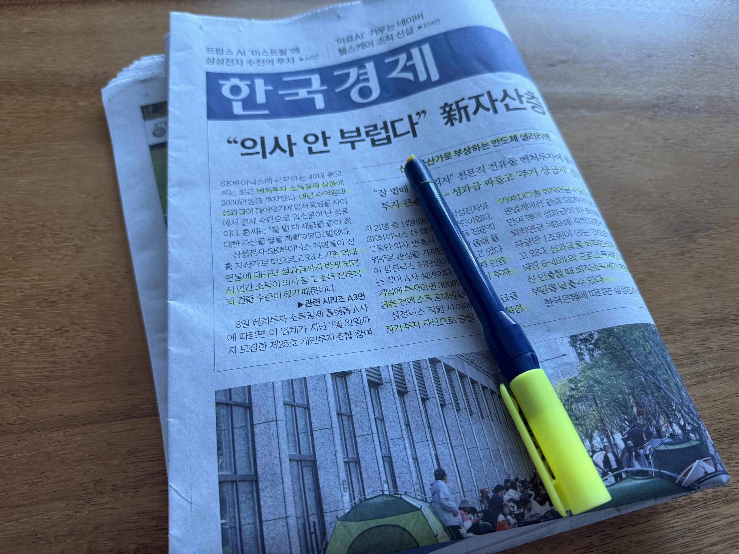 나무 책상 위 한국경제신문 1면과 볼펜·형광펜. 머리기사는 '"의사 안 부럽다" 新자산층'이고 첫 문단 여러 곳에 형광펜이 그어져 있다. 지면 아래에는 건물 앞 인도에 텐트가 늘어선 사진이 있다.